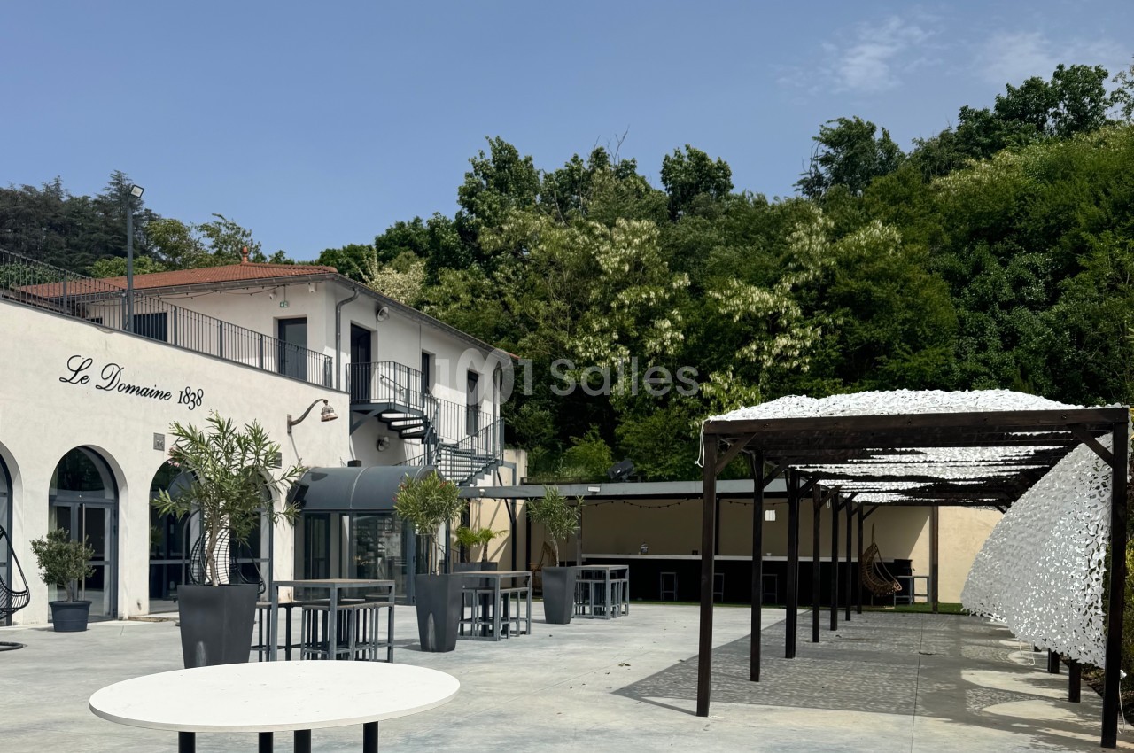 Terrasse extérieure avec tables, pergola moderne et bâtiment blanc entouré de verdure sous un ciel dégagé.