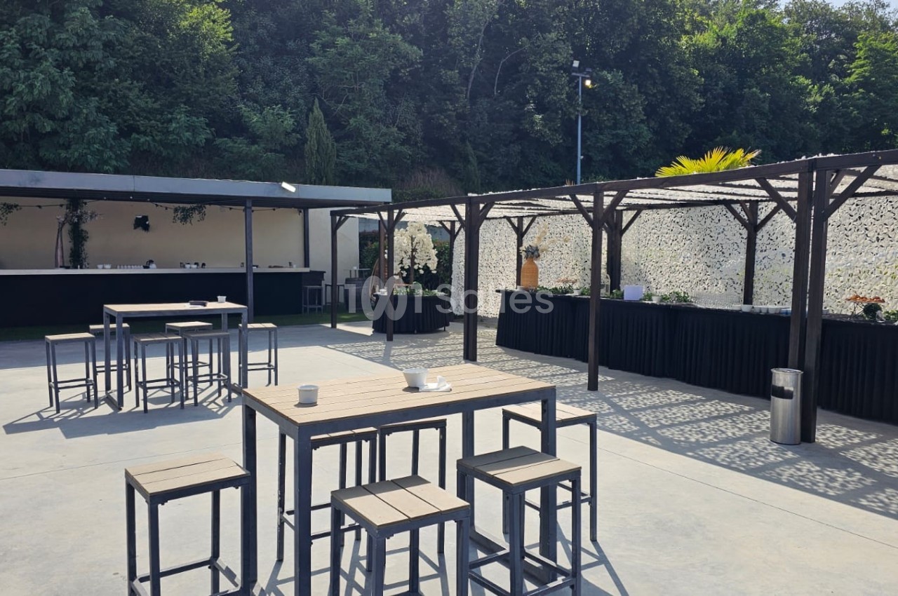 Terrasse extérieure aménagée avec tables hautes, chaises et pergolas, entourée de verdure en arrière-plan.