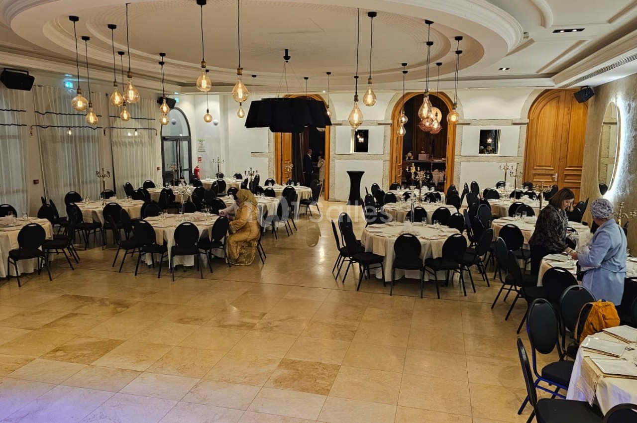 Salle de réception élégante avec tables dressées, chaises noires et éclairage suspendu dans un décor lumineux.