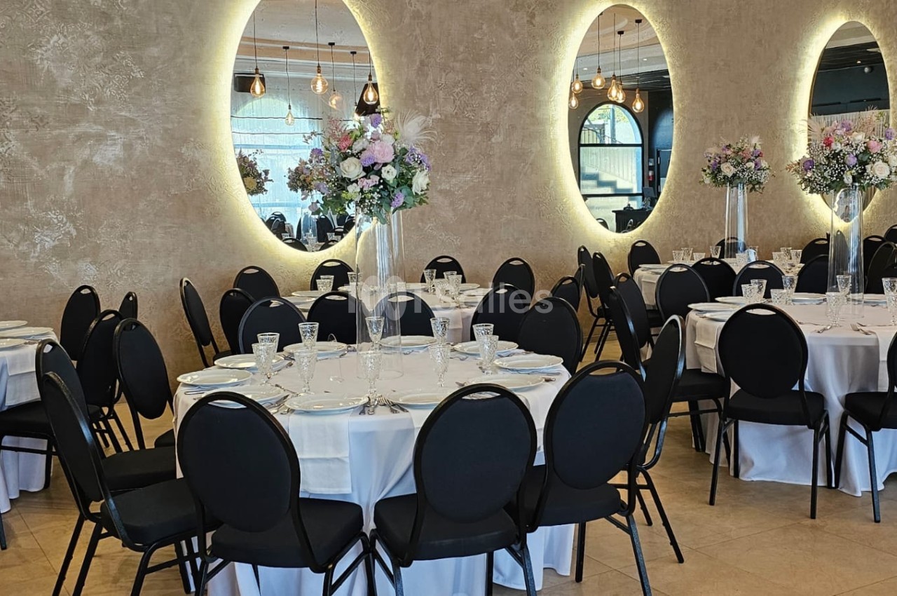 Salle de réception élégante avec tables rondes dressées, chaises noires et décorations florales sur fond de miroirs lumineux.