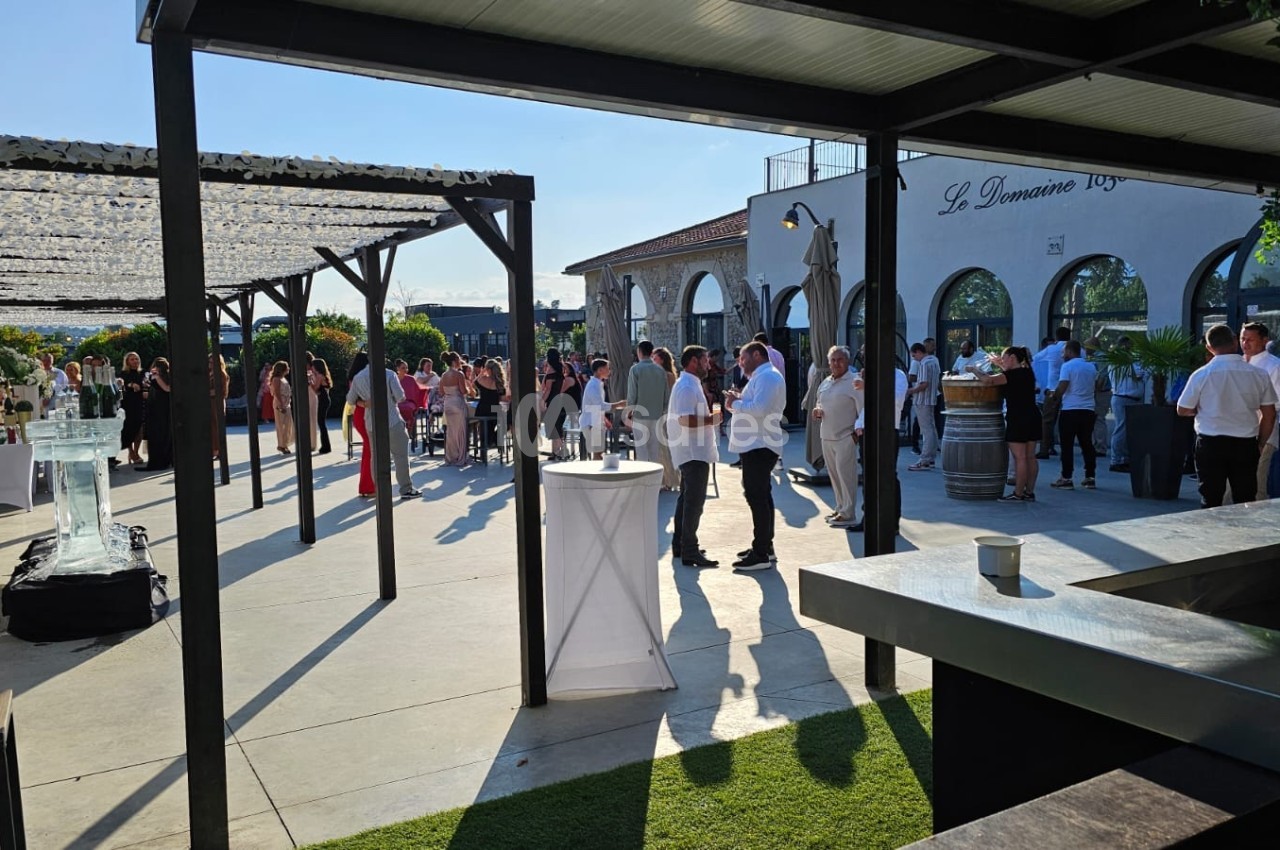 Groupe de personnes rassemblées dans une cour extérieure ensoleillée, entourée de pergolas et de tables hautes.