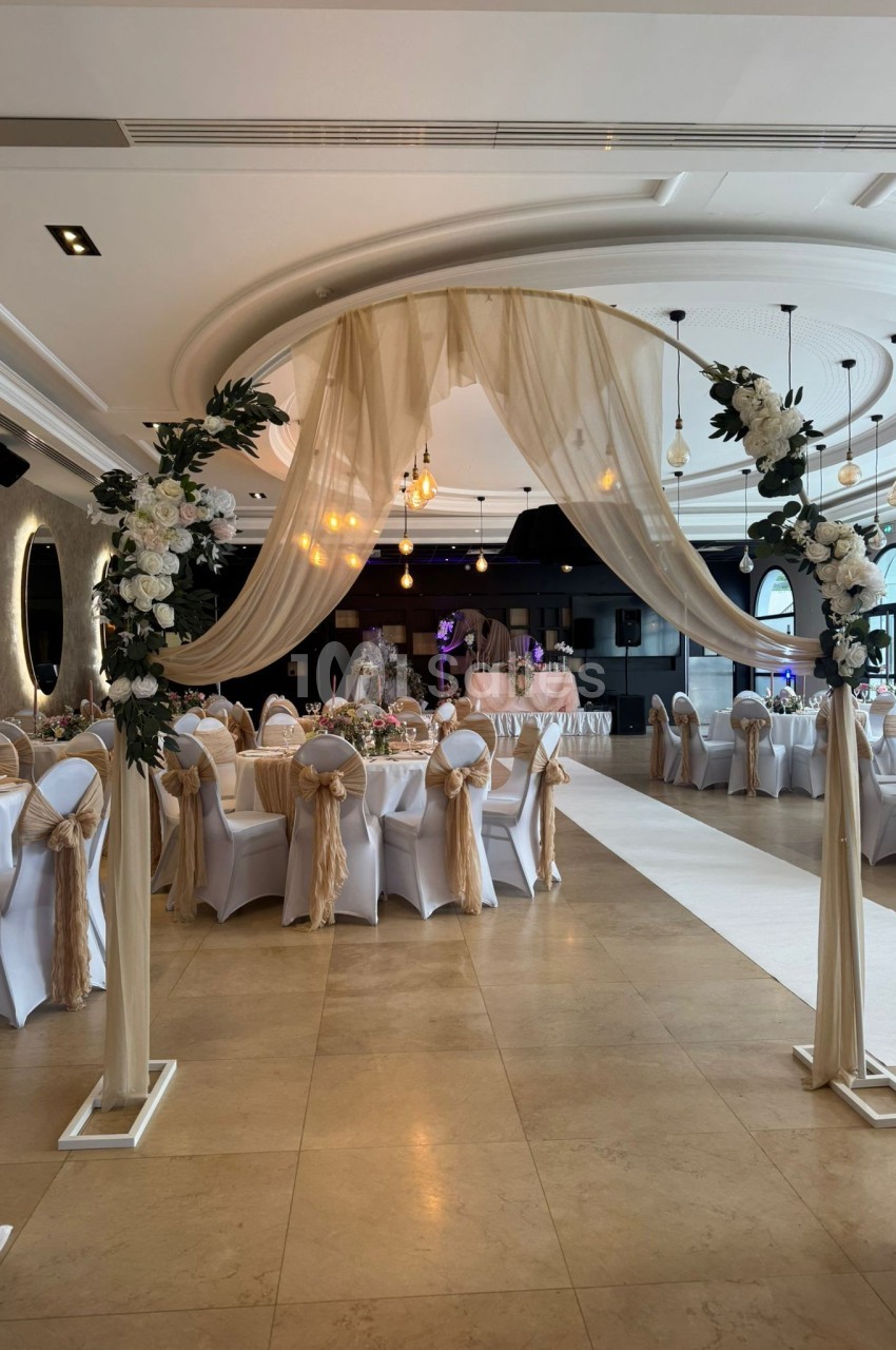 Salle de réception décorée avec des tables dressées, des chaises ornées de nœuds beige et une arche florale en premier plan.