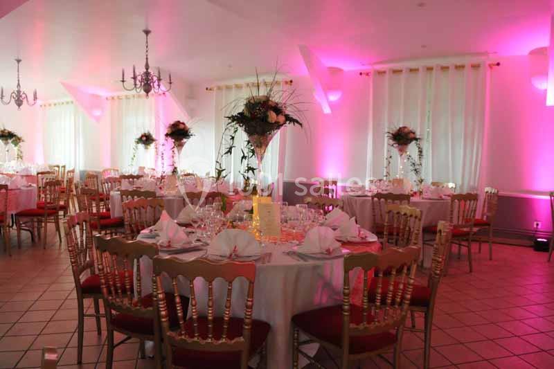 Salle de réception décorée avec des tables rondes, nappes blanches, chaises rouges et éclairage rose.
