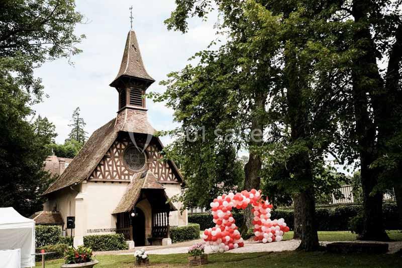 Petite chapelle en bois et pierre entourée d'arbres, avec une arche décorée de ballons rouges et blancs au premier plan.