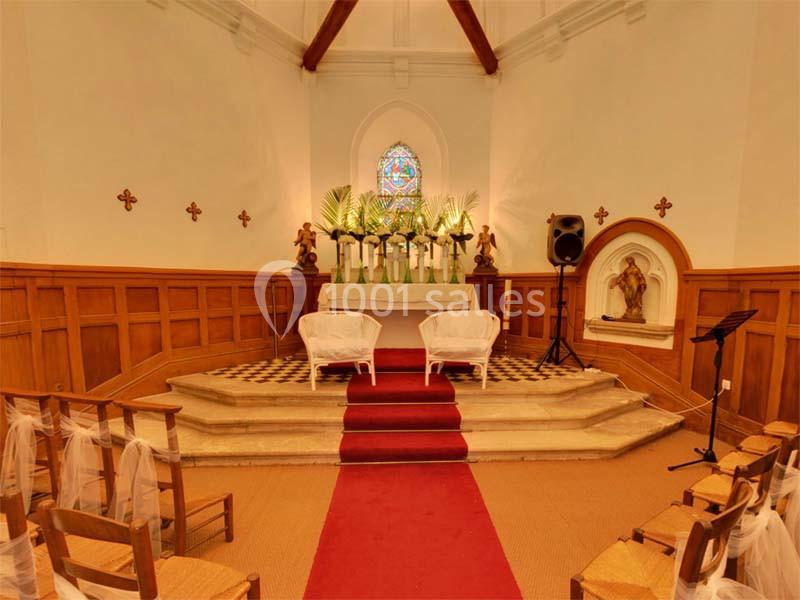 Intérieur d'une chapelle avec autel décoré de plantes, vitrail coloré et chaises alignées sur un tapis rouge.