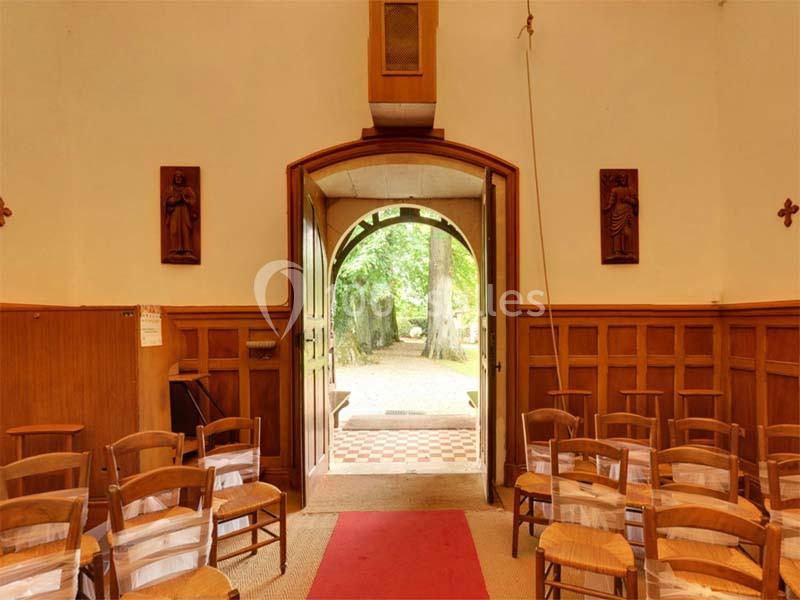 Vue intérieure d'une petite chapelle avec des chaises en bois, une porte ouverte donnant sur un chemin bordé d'arbres.