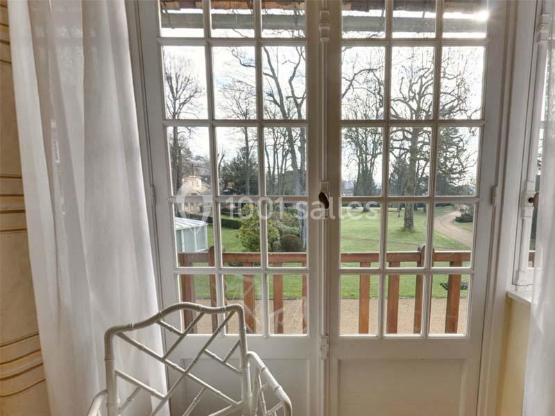 Vue sur un jardin arboré depuis une fenêtre à carreaux, avec une chaise blanche au premier plan.