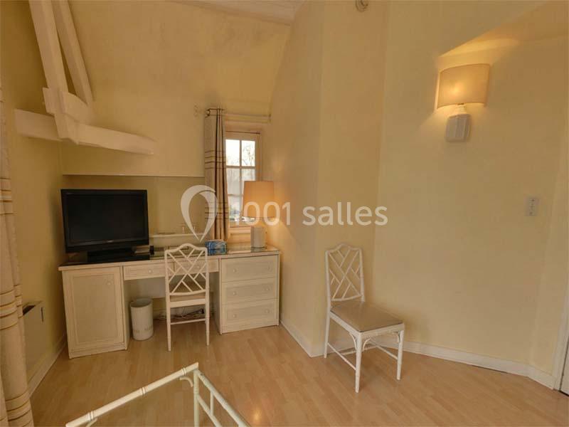 Chambre lumineuse avec bureau, chaise, télévision, lampe et murs beige clair, parquet au sol.