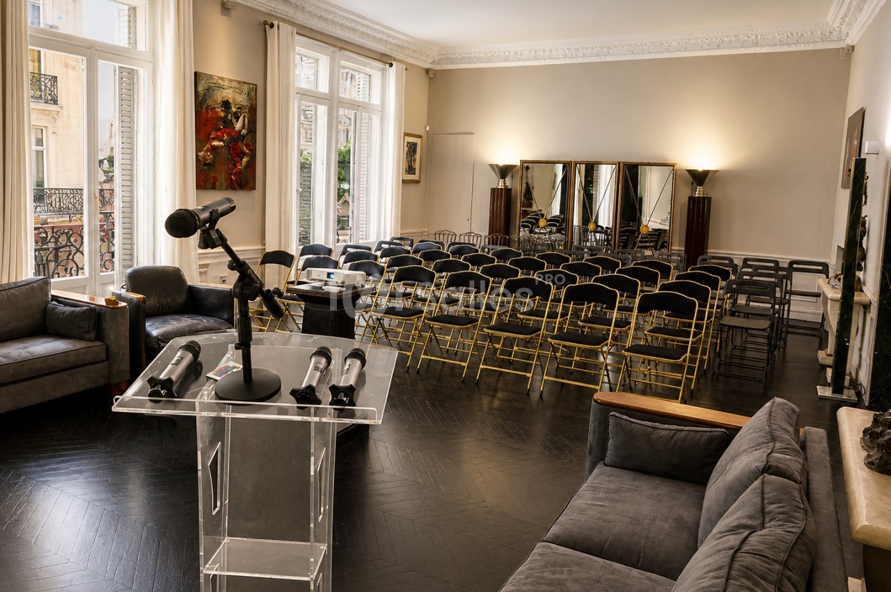 Salle lumineuse avec chaises alignées, pupitre transparent équipé de micros, canapés et grandes fenêtres donnant sur l…