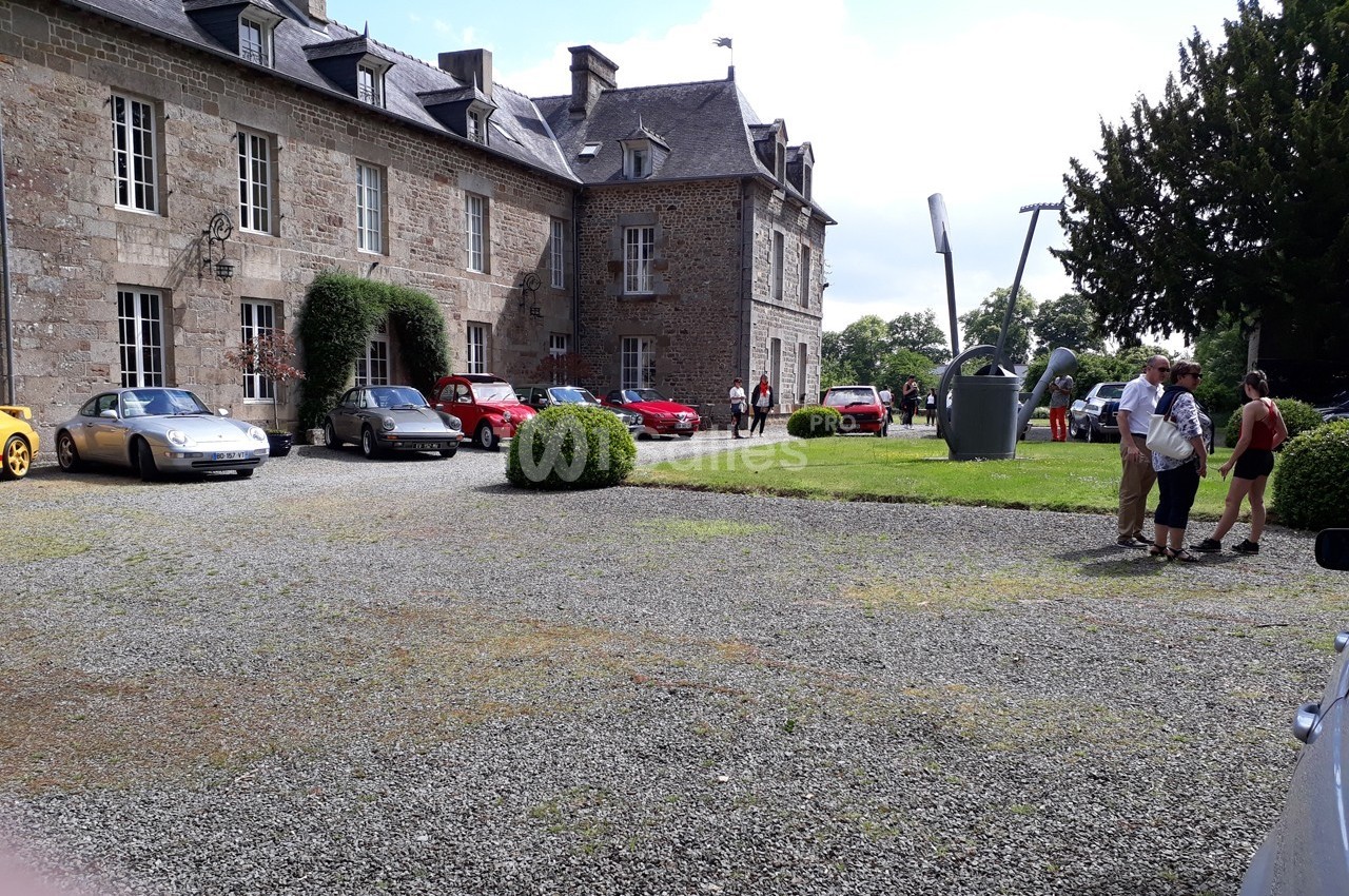 Plusieurs voitures anciennes garées devant un bâtiment en pierre, avec des personnes discutant à proximité.