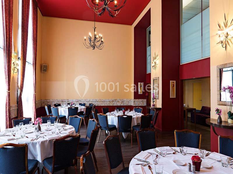 Salle de réception élégante avec tables rondes dressées, chaises noires, hauts plafonds et lustres décoratifs.