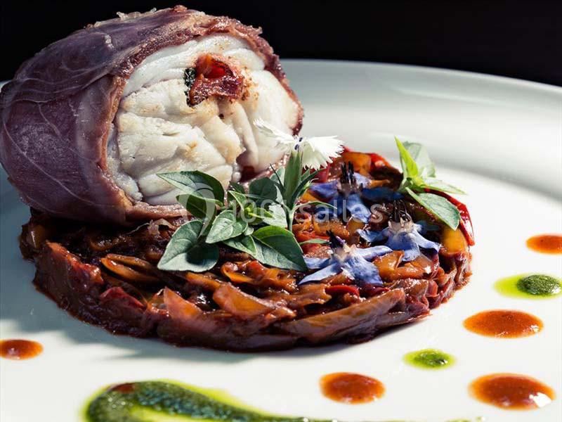 Roulé de poisson entouré de jambon, servi sur un lit de légumes confits avec des herbes et des fleurs comestibles.