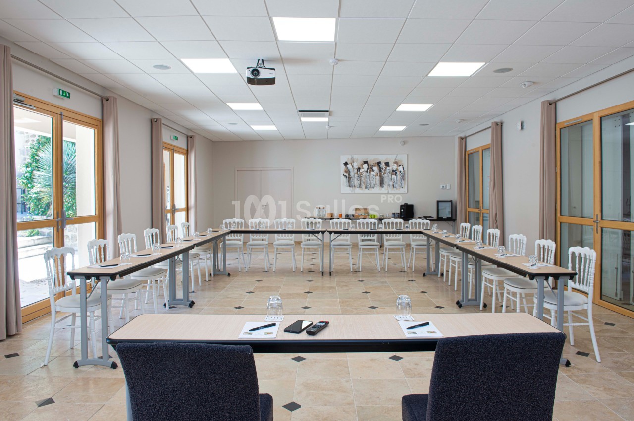 Salle de réunion lumineuse avec tables disposées en U, chaises blanches, projecteur au plafond et grandes baies vitrées.