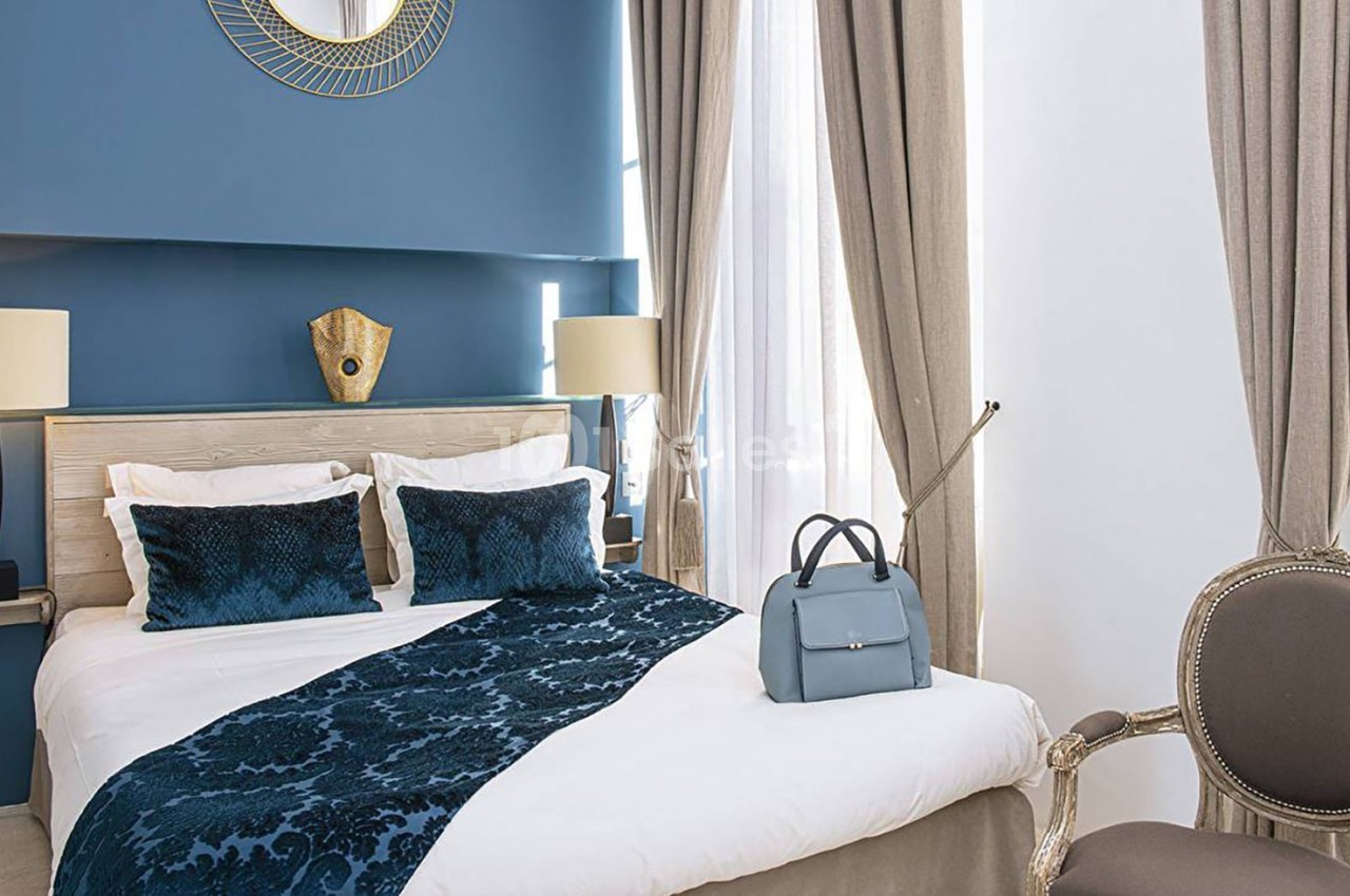 Chambre élégante avec lit double, couvre-lit bleu, rideaux beiges, fauteuil et sac posé sur une chaise.