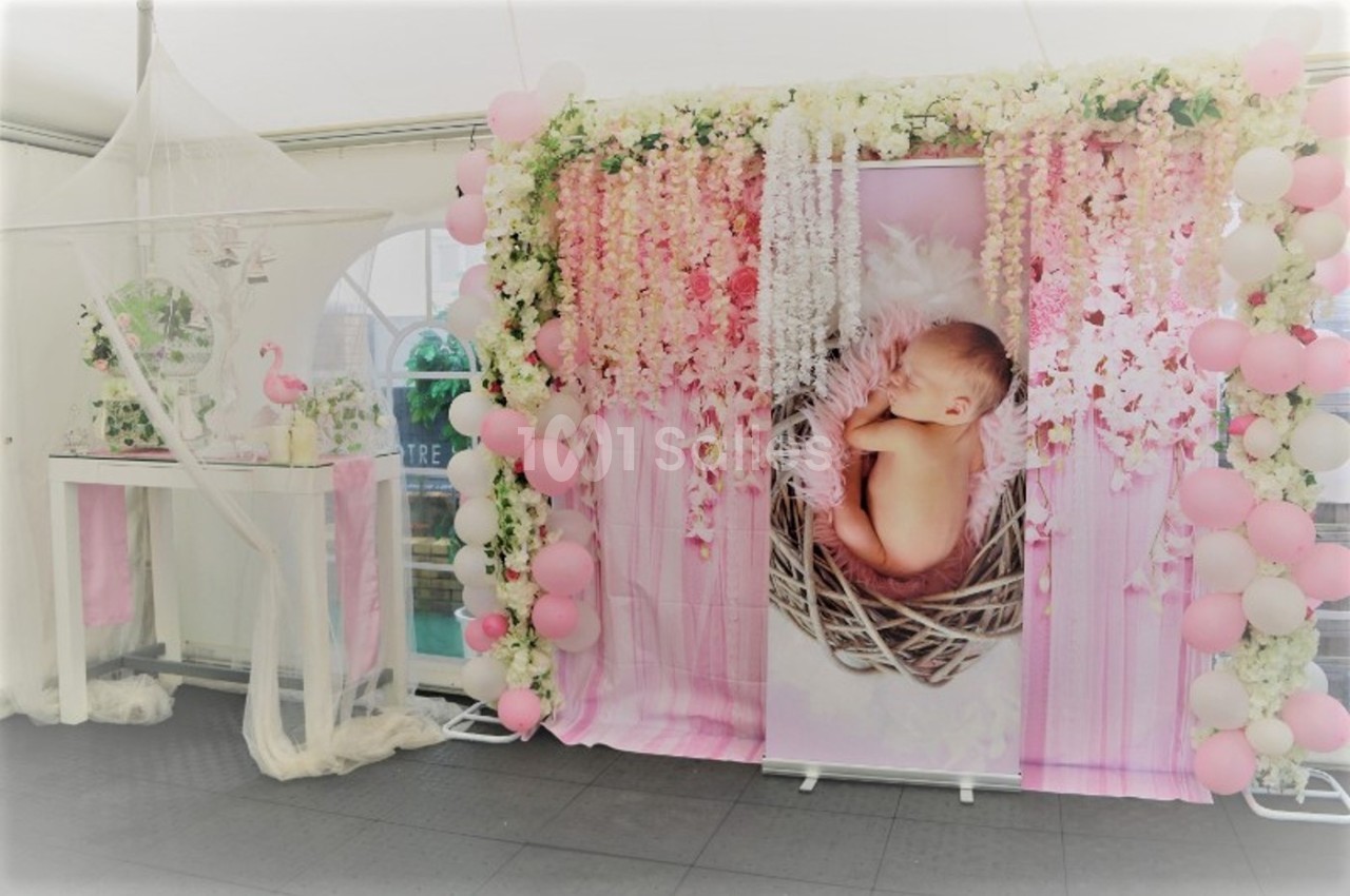 Décoration de fête avec un panneau montrant un bébé dans un nid, entouré de fleurs, ballons roses et voilage blanc.