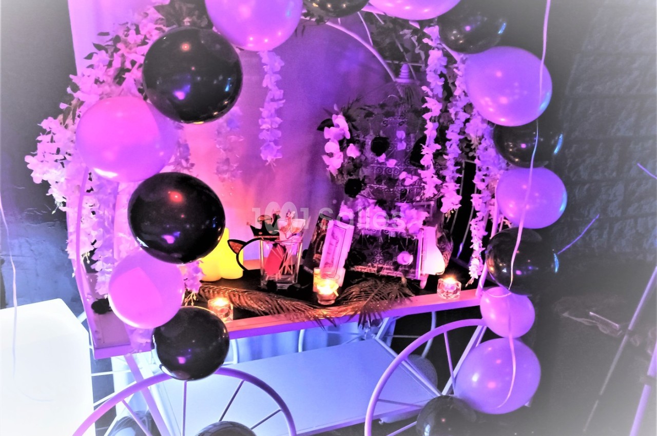 Décoration festive avec ballons noirs et violets, guirlandes florales et bougies sur une table illuminée.