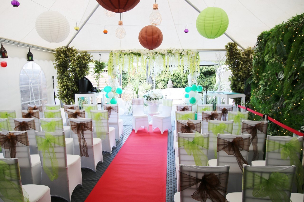 Salle de cérémonie décorée avec des chaises blanches ornées de rubans verts et marron, tapis rouge et lanternes suspendues.