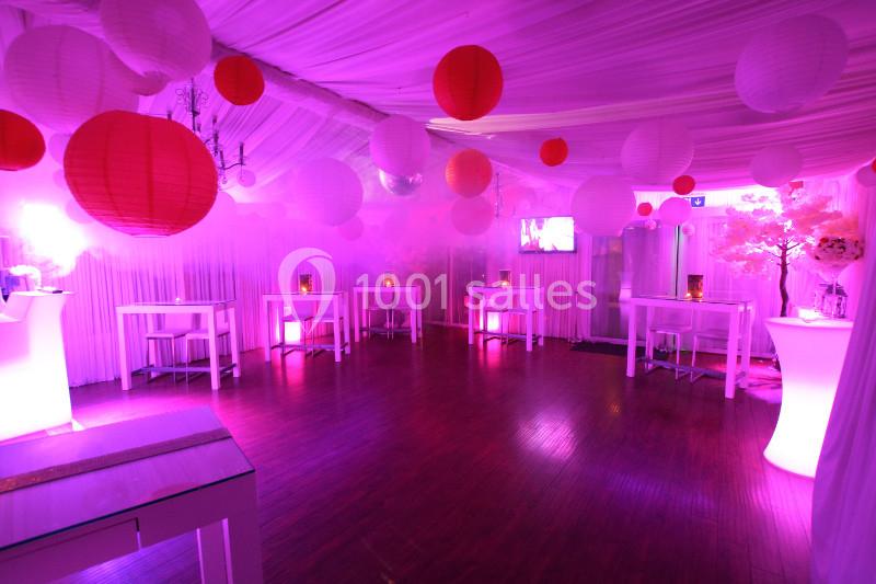 Salle décorée avec des lanternes rouges et blanches, éclairage rose, tables hautes et ambiance festive.