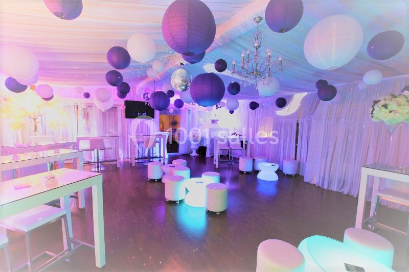Salle décorée pour un événement, avec lanternes suspendues, tables hautes, tabourets blancs et éclairage coloré.