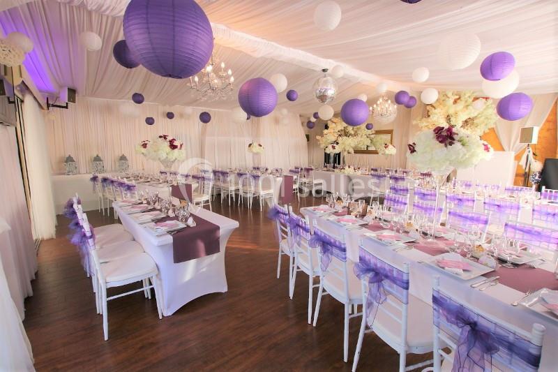 Salle de réception décorée avec des lanternes violettes, des nappes blanches et des compositions florales élégantes.