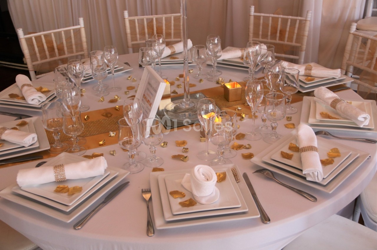Table ronde élégamment dressée avec vaisselle blanche, serviettes pliées, décorations dorées et bougies.