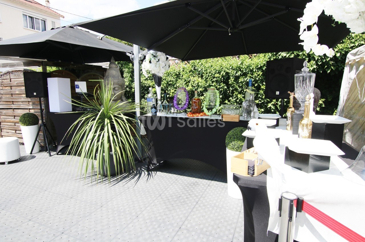 Bar extérieur avec comptoir noir, décorations florales, plantes, fontaines à boissons et parasols noirs.