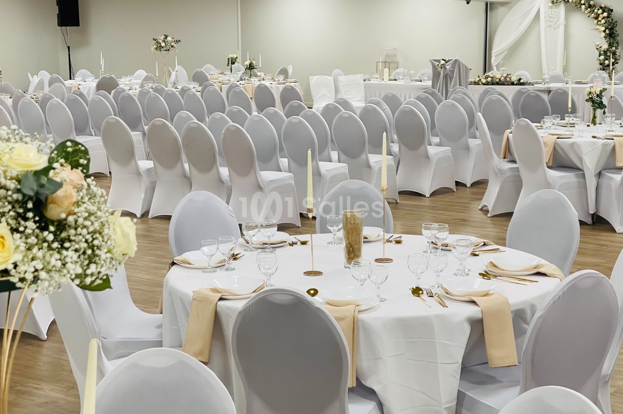 Salle de réception décorée pour un événement, avec des tables rondes dressées, nappes blanches et chaises couvertes.