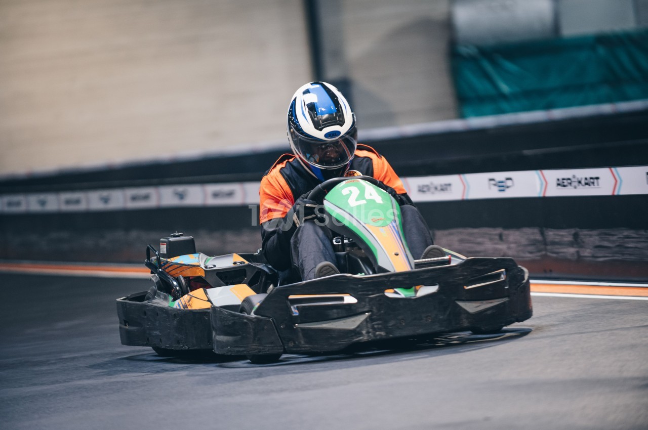 Un pilote en combinaison orange conduit un kart sur une piste intérieure.