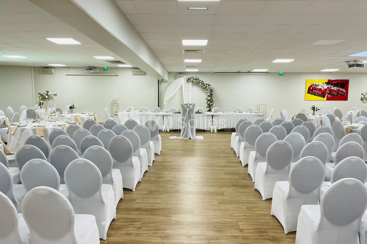 Salle de réception décorée pour un événement, avec des chaises blanches alignées et une arche florale au fond.