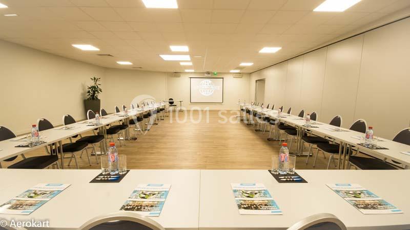 Salle de réunion lumineuse avec tables en U, chaises, bouteilles d'eau et brochures, écran de projection en fond.