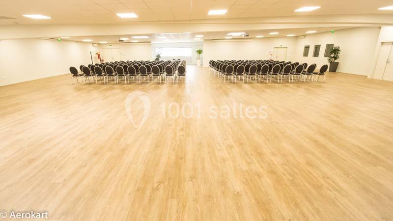 Salle spacieuse avec parquet clair, rangées de chaises noires alignées face à un écran blanc, éclairage naturel.