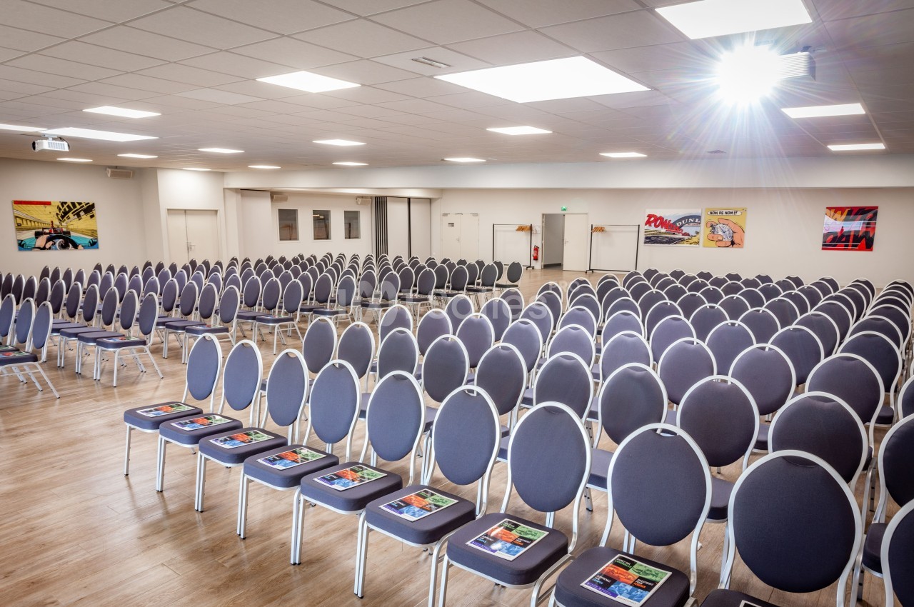 Salle de conférence lumineuse avec des rangées de chaises grises alignées, chacune équipée d'un document coloré.