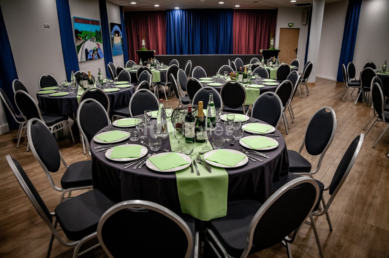 Salle de réception avec tables rondes dressées, nappes noires, serviettes vertes et bouteilles disposées.