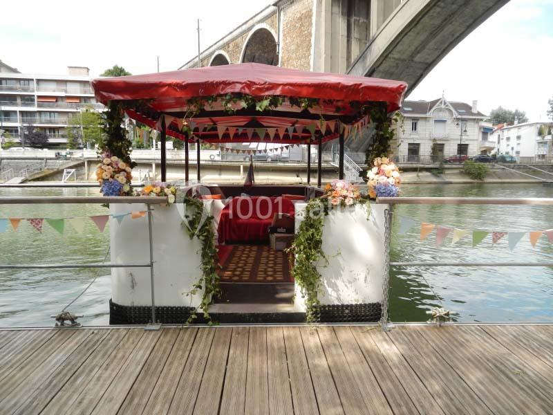 Bateau décoré de fleurs et guirlandes colorées, amarré sur une rivière près d'un pont en pierre.