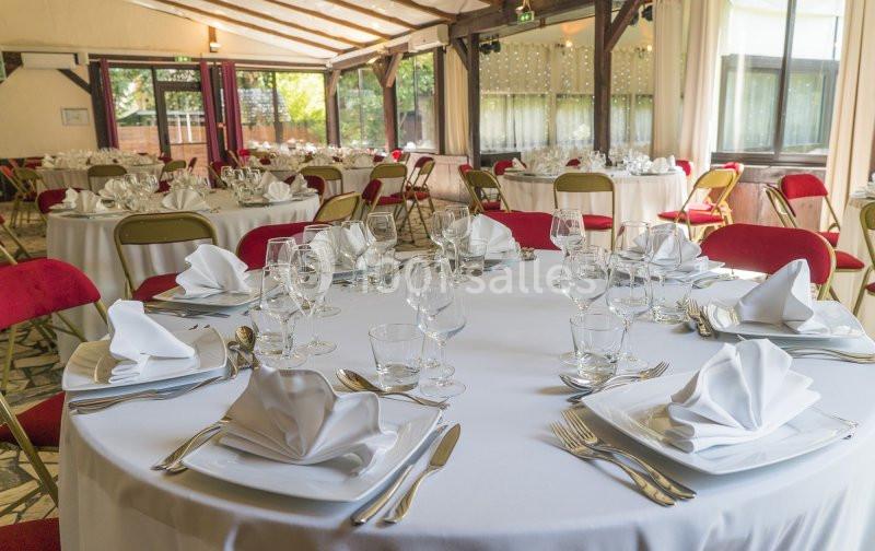Salle de réception avec des tables rondes dressées, nappes blanches, serviettes pliées et chaises rouges.