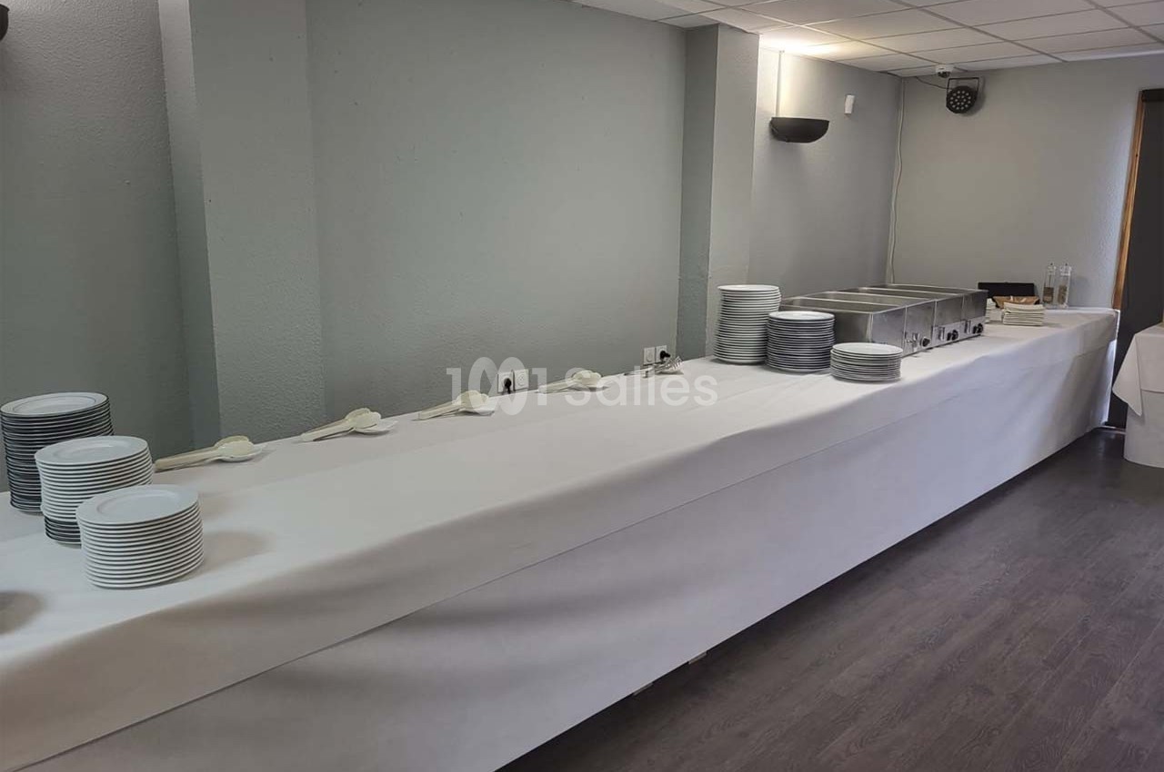 Table de buffet vide avec assiettes empilées, cuillères et chafing dishes dans une salle sobrement décorée.