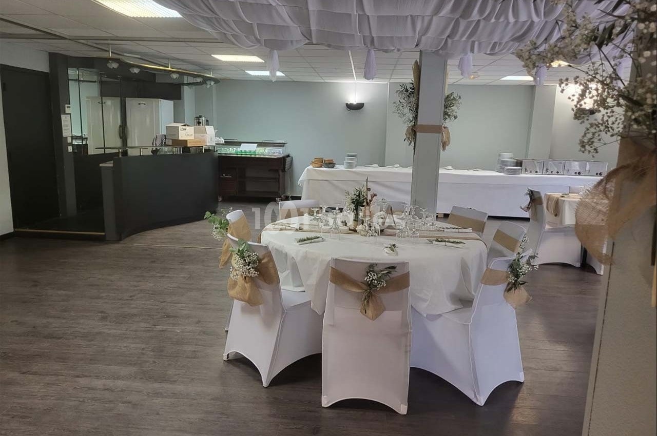 Salle de réception décorée avec des tables rondes, nappes blanches et chaises ornées de rubans et fleurs.