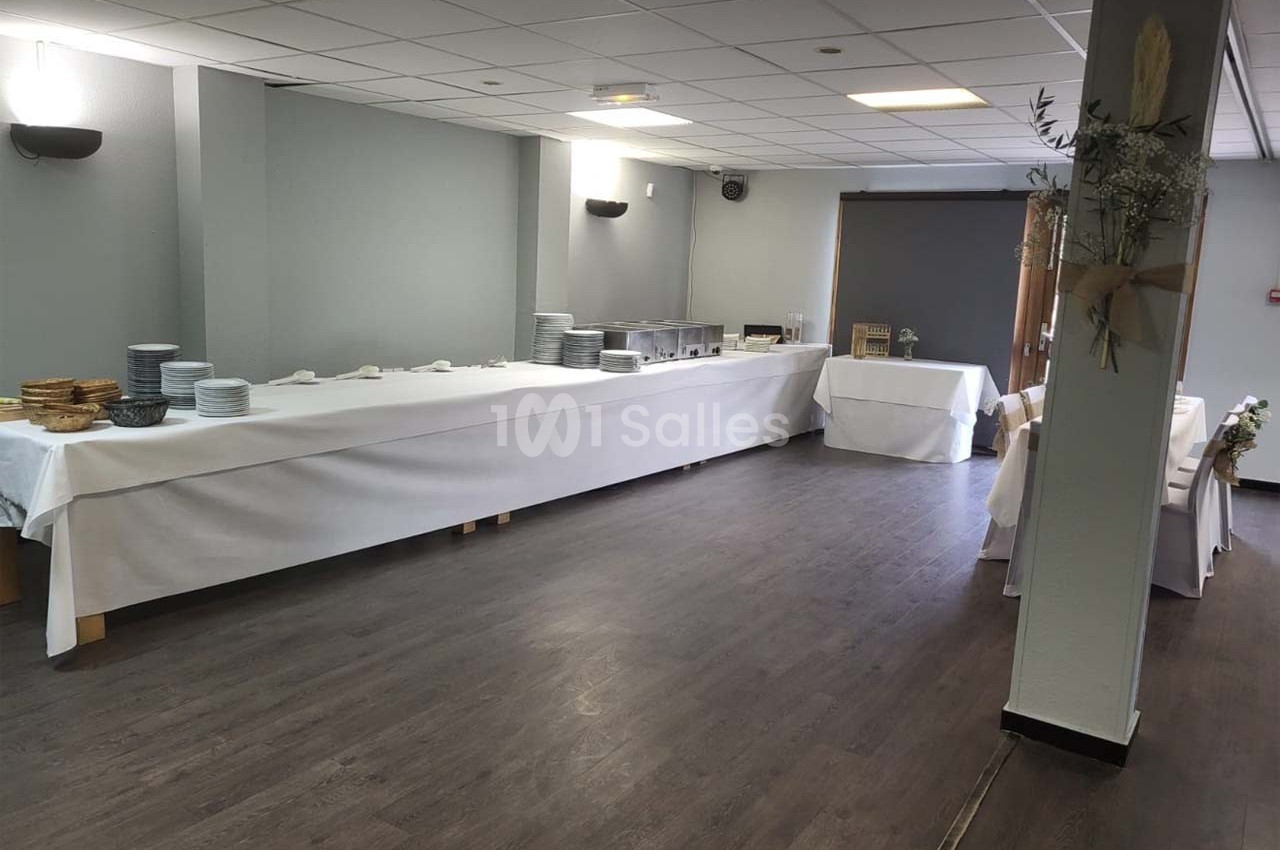 Salle avec tables alignées couvertes de nappes blanches, préparées pour un buffet avec assiettes et plats disposés.