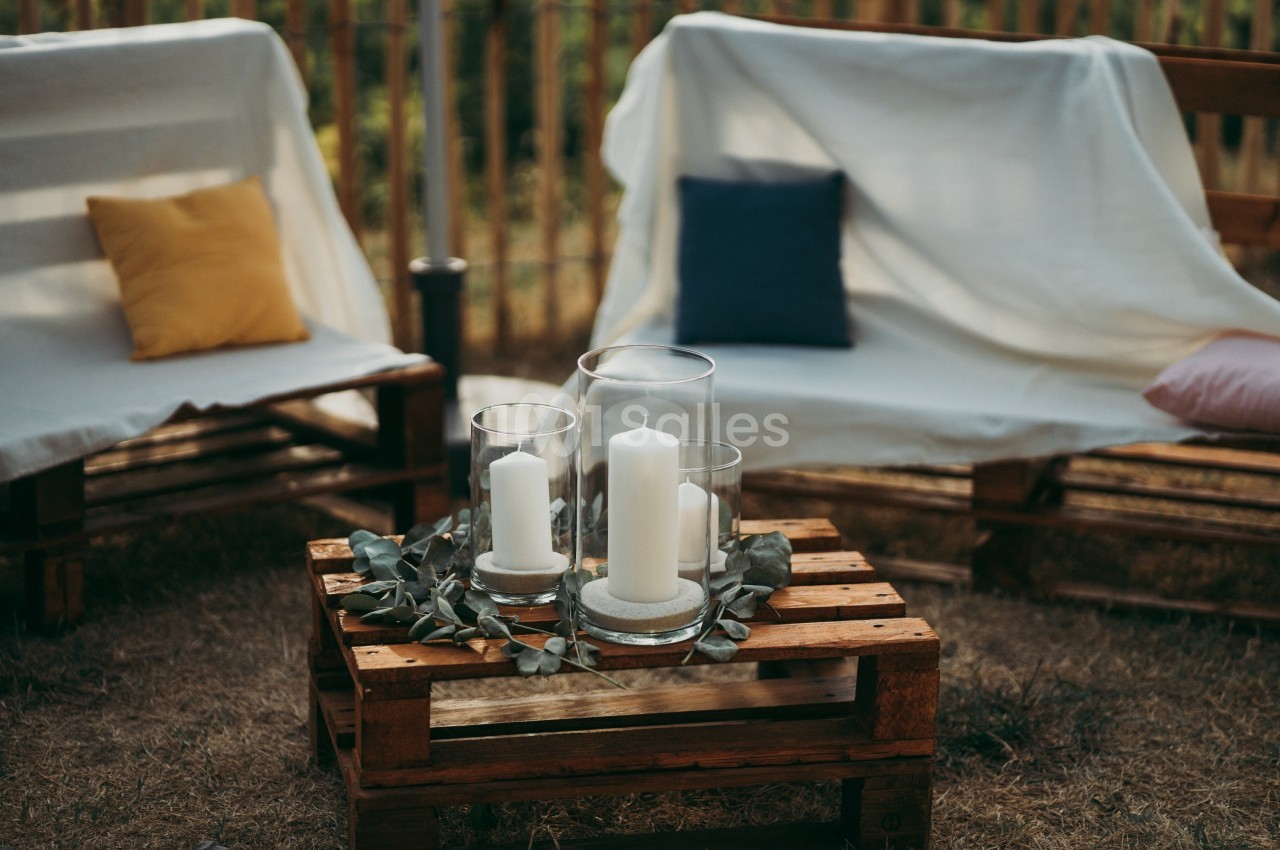 Bougies blanches dans des photophores en verre sur une table en palettes, entourées de feuilles, avec des fauteuils en…