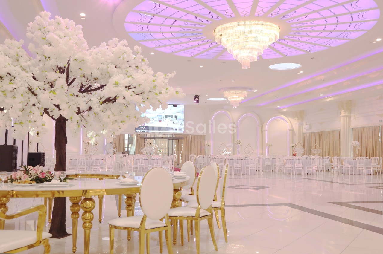 Salle de réception élégante avec plafond lumineux, lustre imposant, table dorée et arbre décoratif aux fleurs blanches.