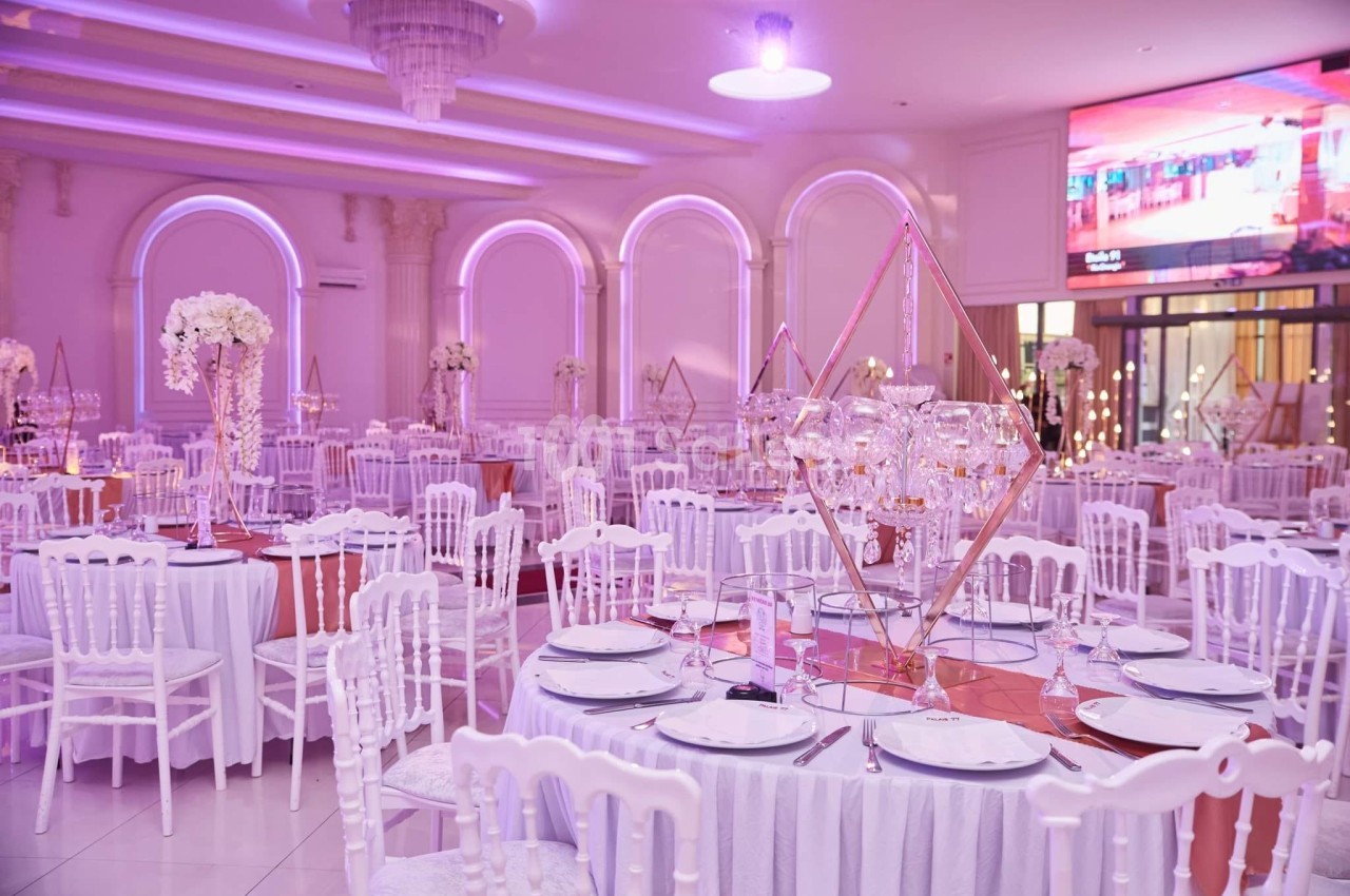 Salle de réception élégante avec tables décorées de nappes blanches, centres de table floraux et éclairage rose.