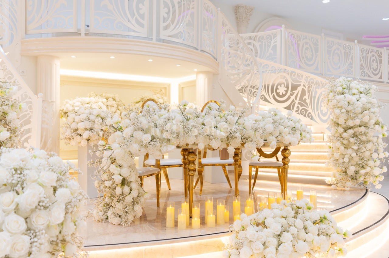 Salle de réception décorée de fleurs blanches, avec une table dorée et des bougies alignées sur des marches illuminées.