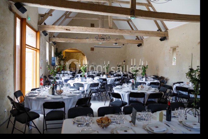 Salle de réception avec des tables rondes dressées, nappes blanches, chandeliers et poutres apparentes au plafond.