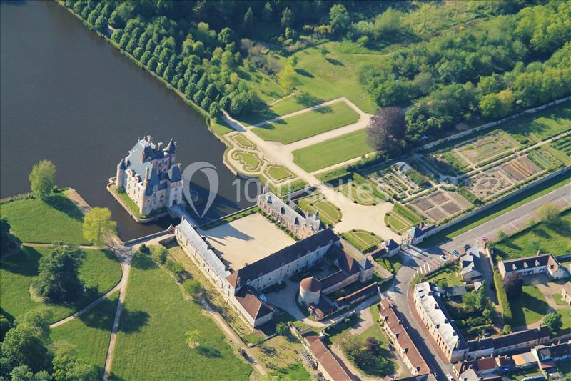 Vue aérienne d'un château entouré d'un jardin structuré, d'un plan d'eau et de bâtiments annexes dans un paysage verdoyant.