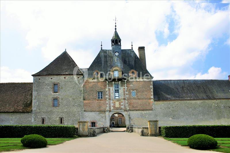 Façade d'un bâtiment historique en pierre et briques avec une tour centrale, entouré de haies et d'un ciel dégagé.