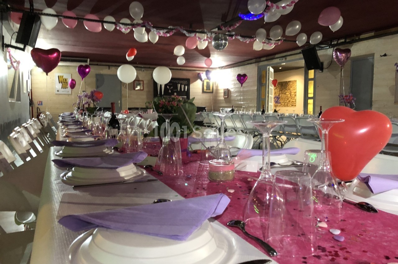 Salle décorée pour une fête avec des ballons suspendus, une table dressée avec nappes roses et serviettes violettes.