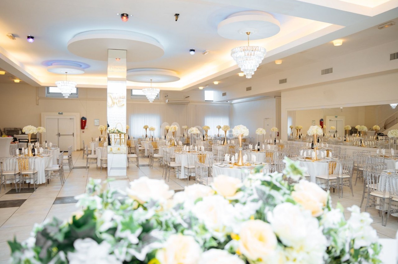 Salle de réception lumineuse décorée de fleurs blanches, tables rondes dressées et lustres suspendus.