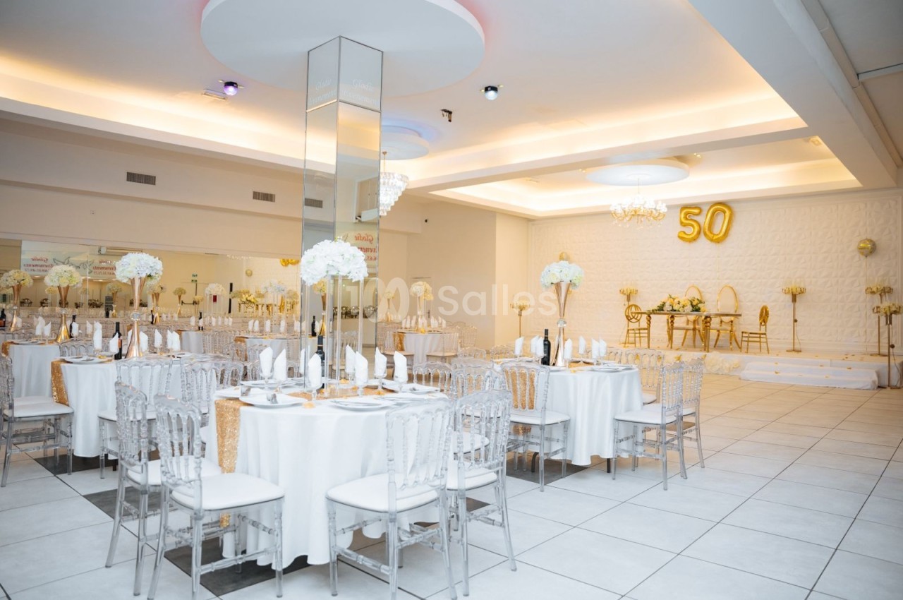 Salle de réception élégante décorée en blanc et or, avec tables dressées et ballon ’50’ en arrière-plan.