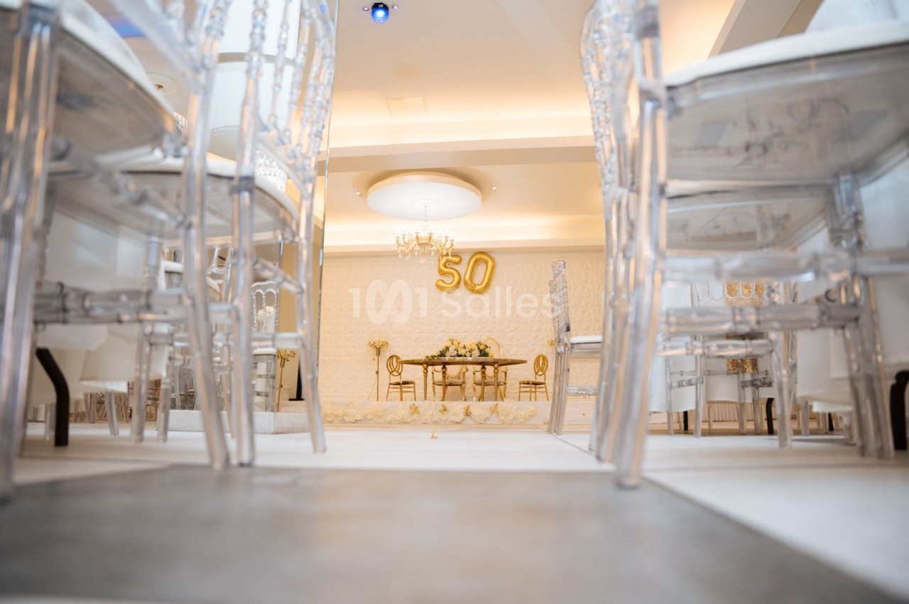 Salle décorée pour une célébration avec des chaises transparentes, une table ornée et le chiffre 50 en ballons dorés.