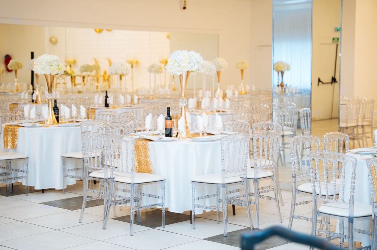 Salle de réception élégante avec tables dressées, nappes blanches, chaises transparentes et centres de table fleuris.